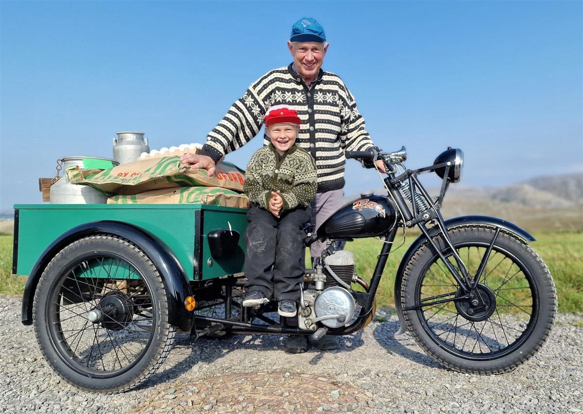 Foto av gammel motorsykkel med lasteplan.  Sjåfør og liten gutt i strikkejakker og caps.  - Klikk for stort bilete