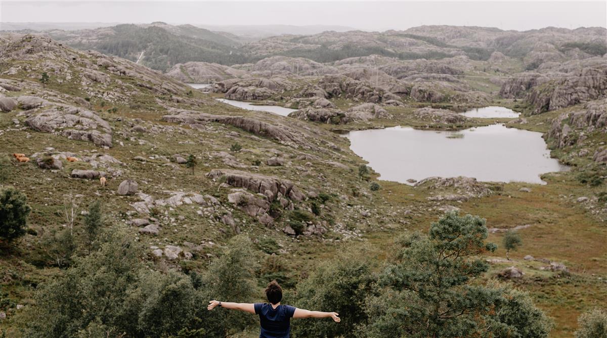 Person med armane utstrekte framfor eit landskap med fjell, vatn og vegetasjon. - Klikk for stort bilete
